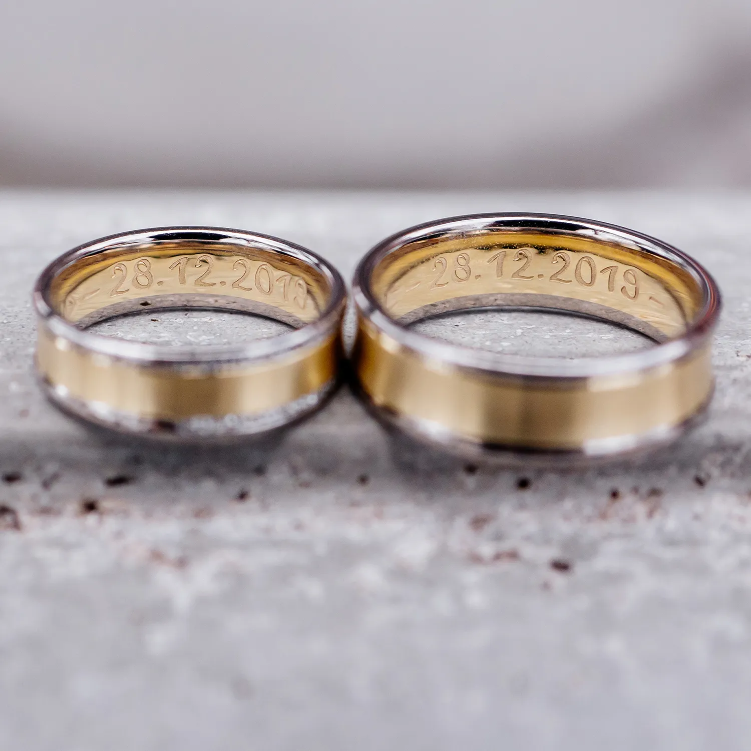 Two gold and silver rings engraved with the date 28.12.2019 on a textured surface.