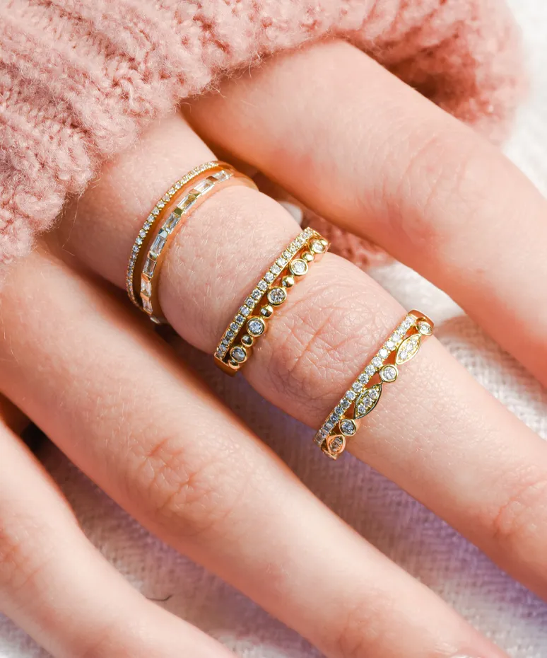 Three elegant gold rings with diamonds, worn on a hand with a soft pink sweater. Shop at Genesis Jewelry in Muscle Shoals.