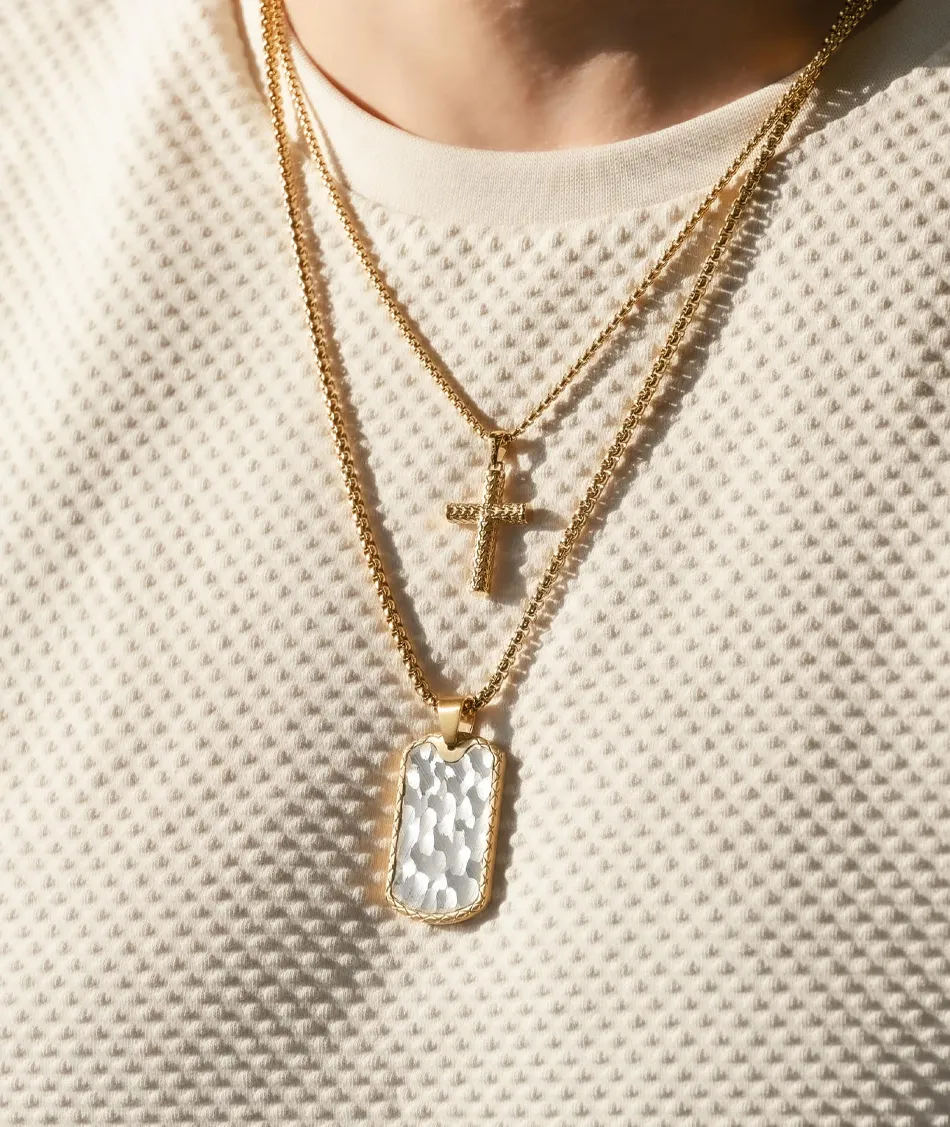 Gold layered necklaces featuring a cross and a textured pendant, worn over a light-colored textured shirt.