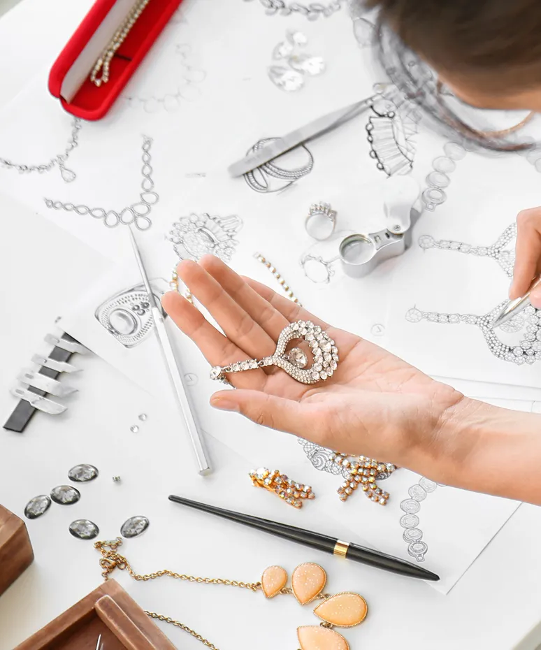 Hand holding a sparkling jewelry piece over design sketches and tools, showcasing custom jewelry creation at Genesis Jewelry.