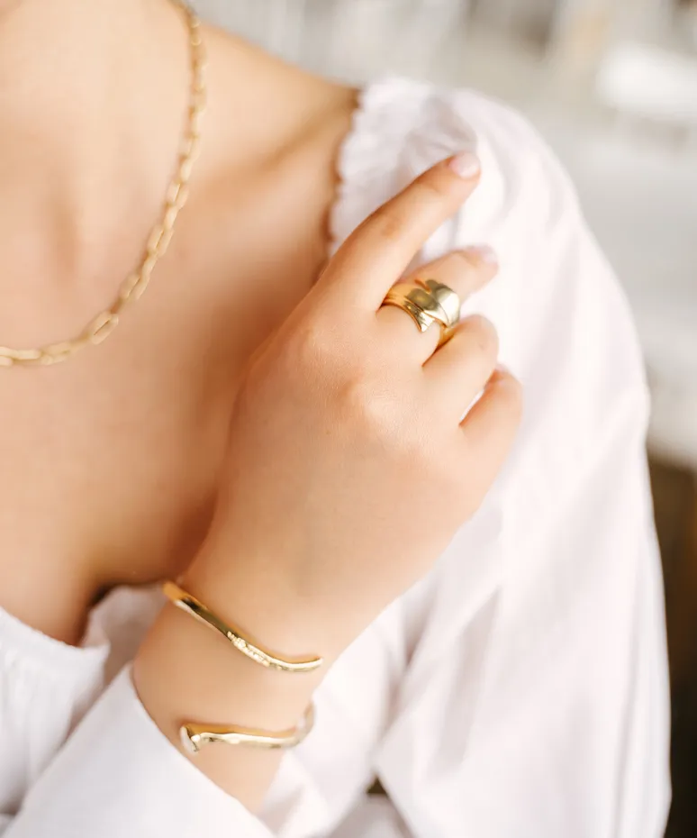 Close-up of a hand wearing gold jewelry: a chain necklace, a ring, and a bracelet, against a soft background.
