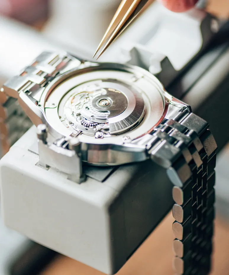 A watch repair technician uses tweezers to work on a watch movement inside a workshop.