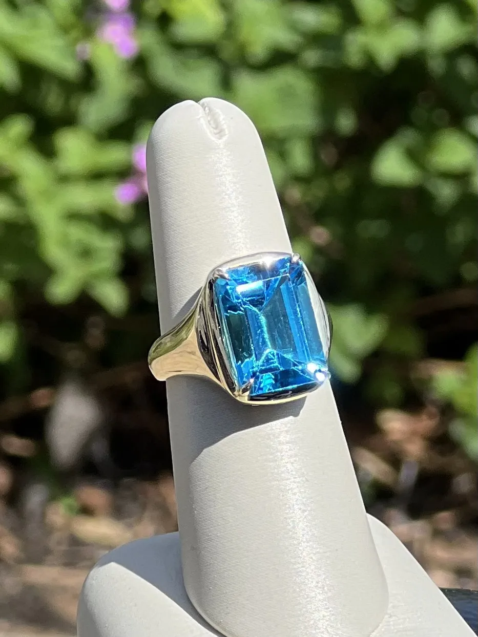 Blue Topaz Emerald Cut 14kt yellow gold ring on ring display in front of flower bush