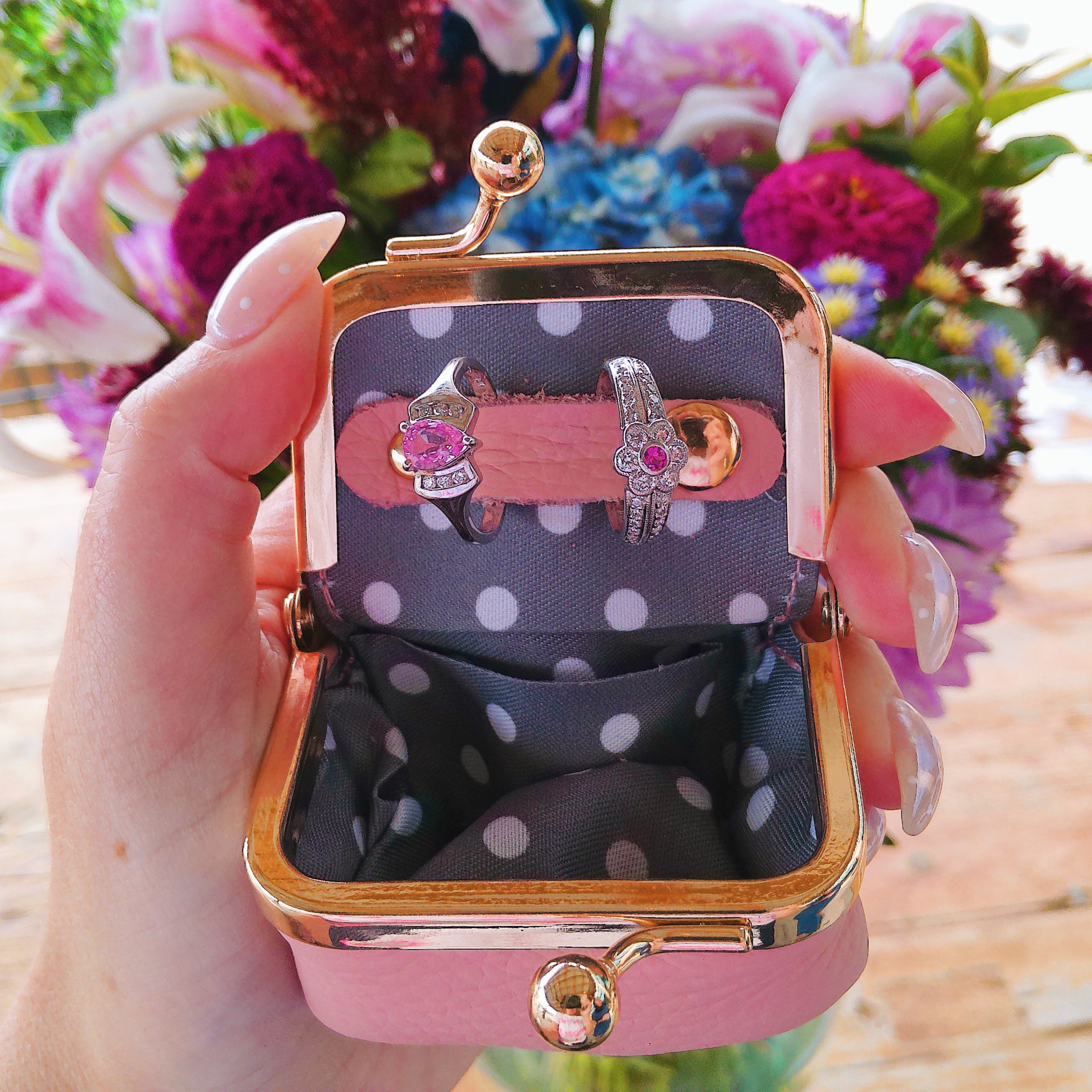 A hand holding a pink jewelry box with two rings inside, surrounded by colorful flowers. Click to explore more jewelry options.