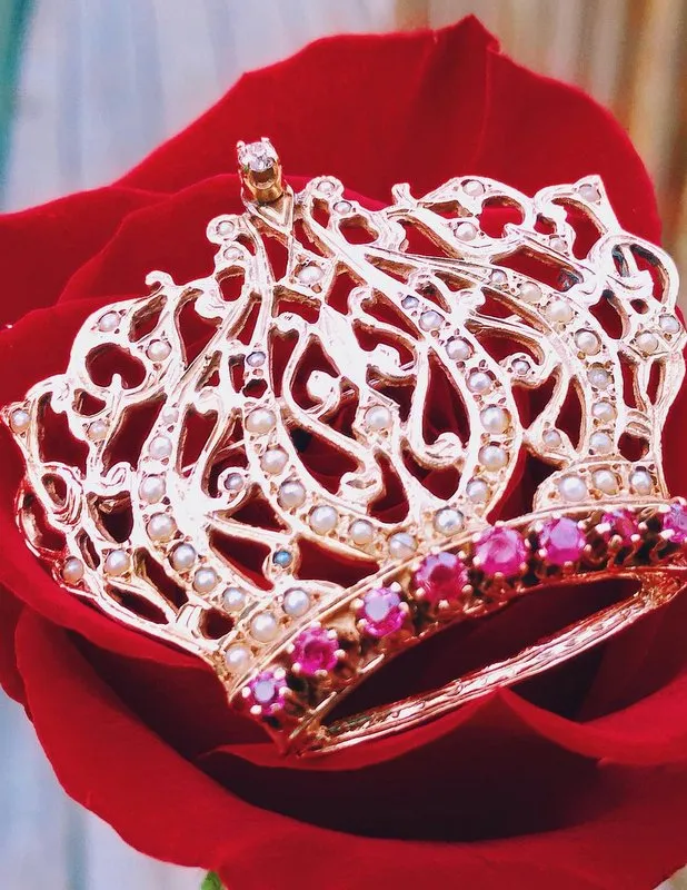 A rose holds a delicate crown adorned with pink & pearl gemstones, symbolizing elegance and beauty. Explore pins and brooches