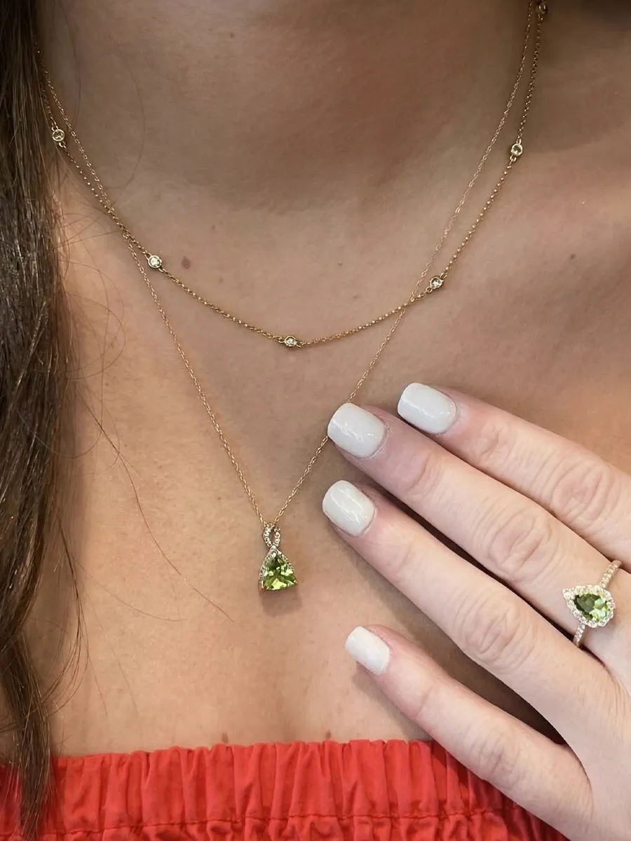 A person wearing a layered gold necklace with a green gemstone pendant and matching ring, with white nail polish.