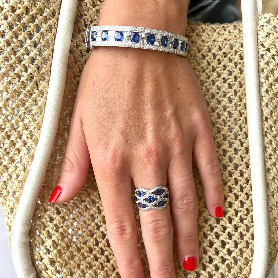 Hand with red nails wearing a sapphire bracelet and ring, holding a woven bag.