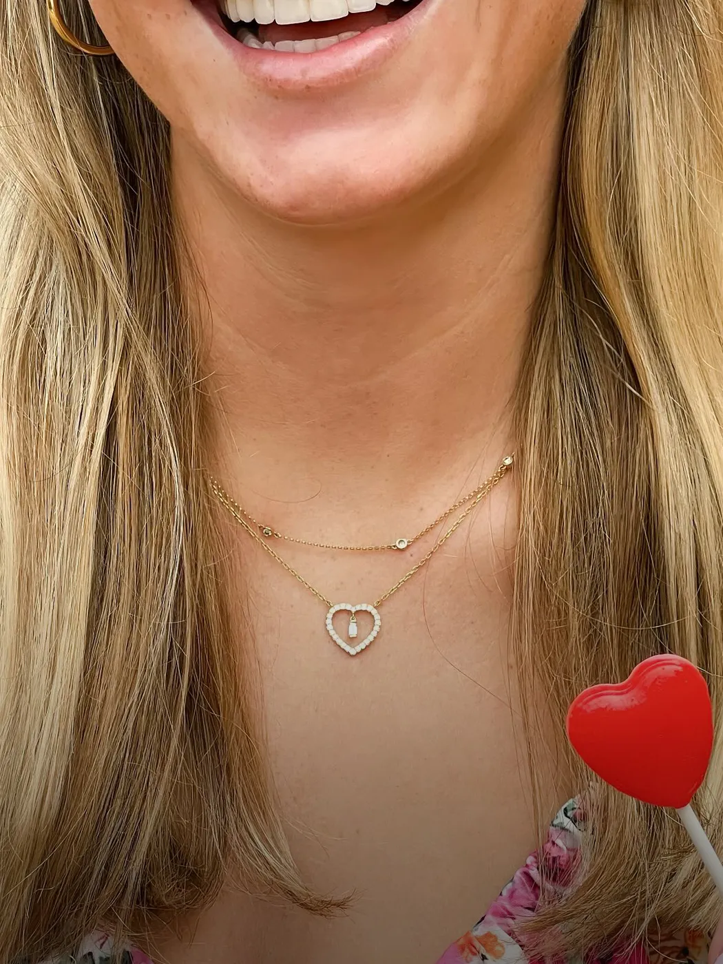 Smiling woman wearing a heart-shaped necklace, holding a heart lollipop.