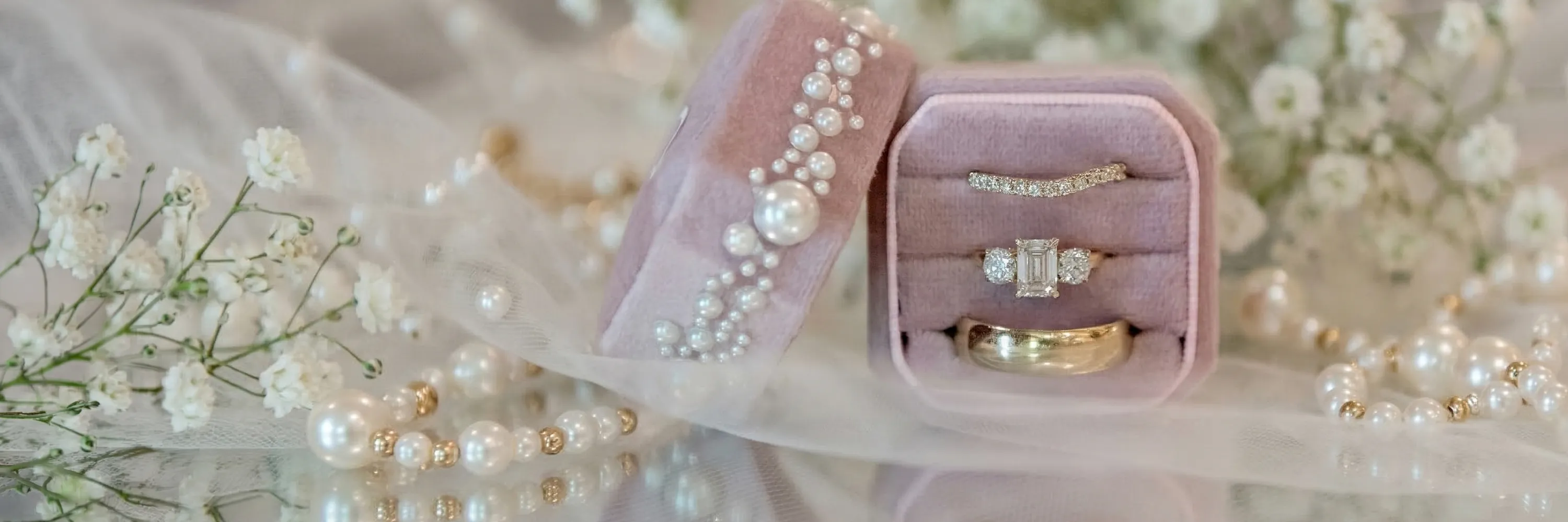 Pink velvet ring box with diamond rings and pearls, surrounded by white flowers.