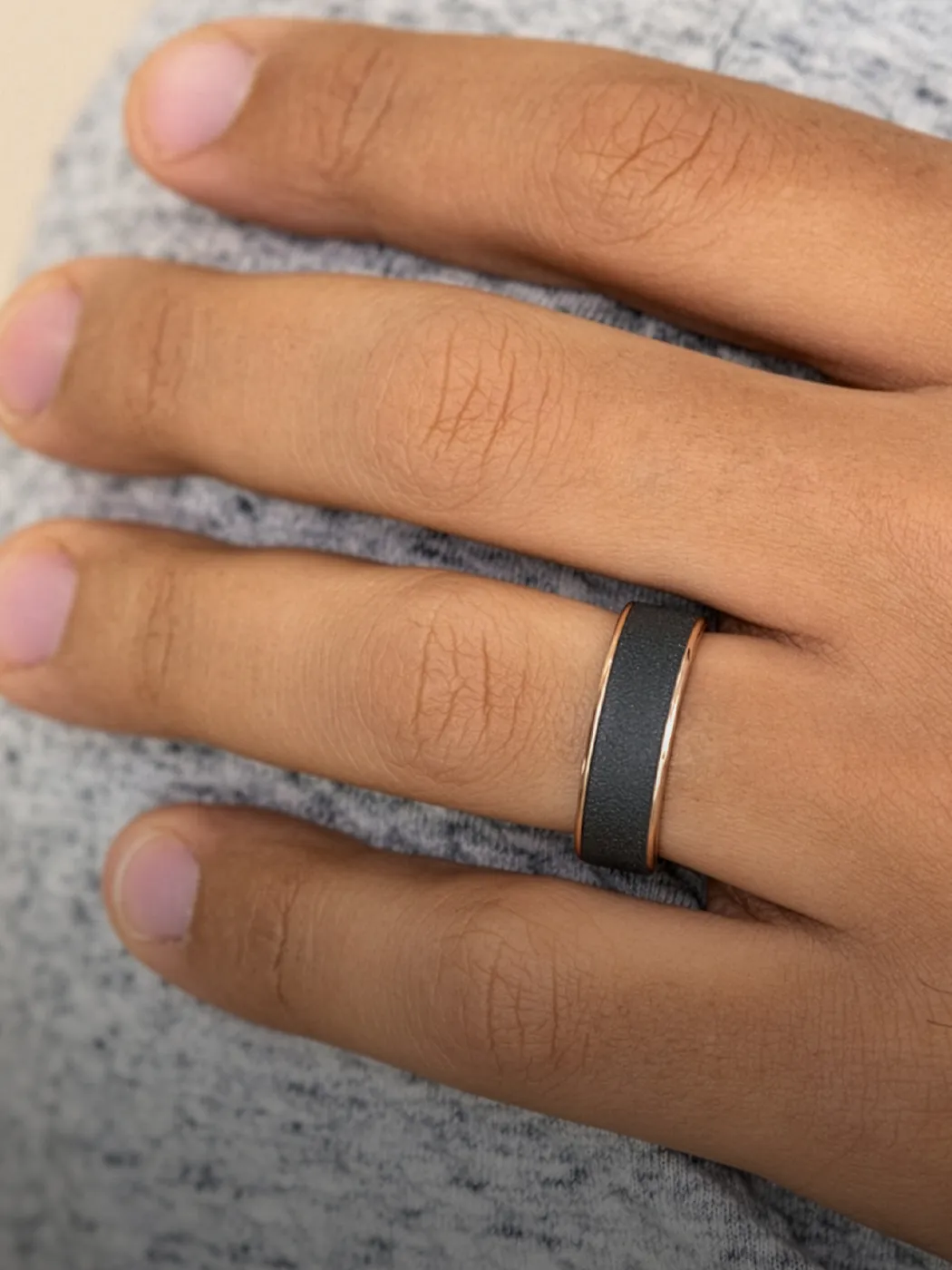 A hand wearing a sleek black men's band ring on a gray fabric background.