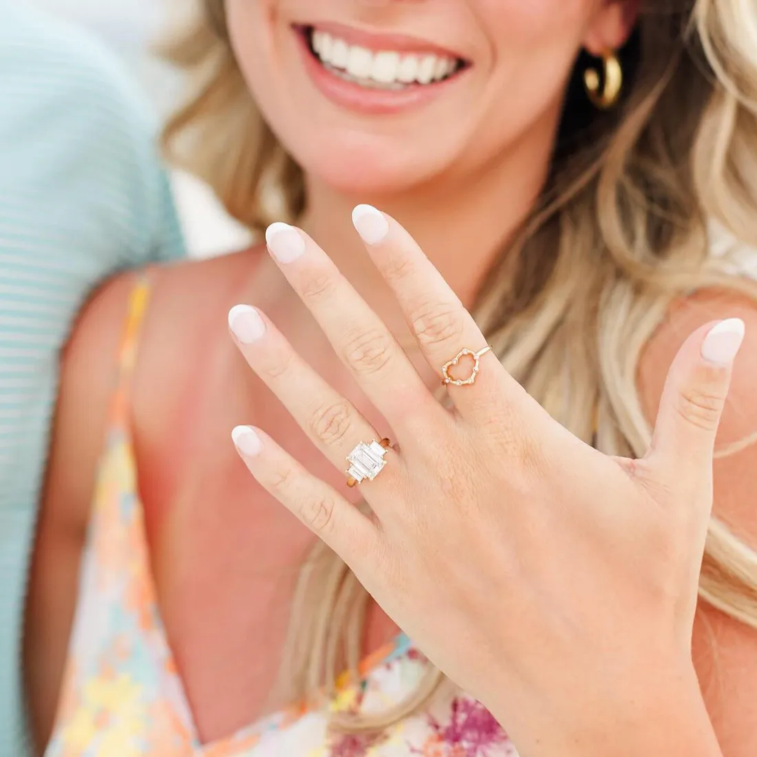 A woman smiling, showing her hand with two rings, one featuring a large gemstone.