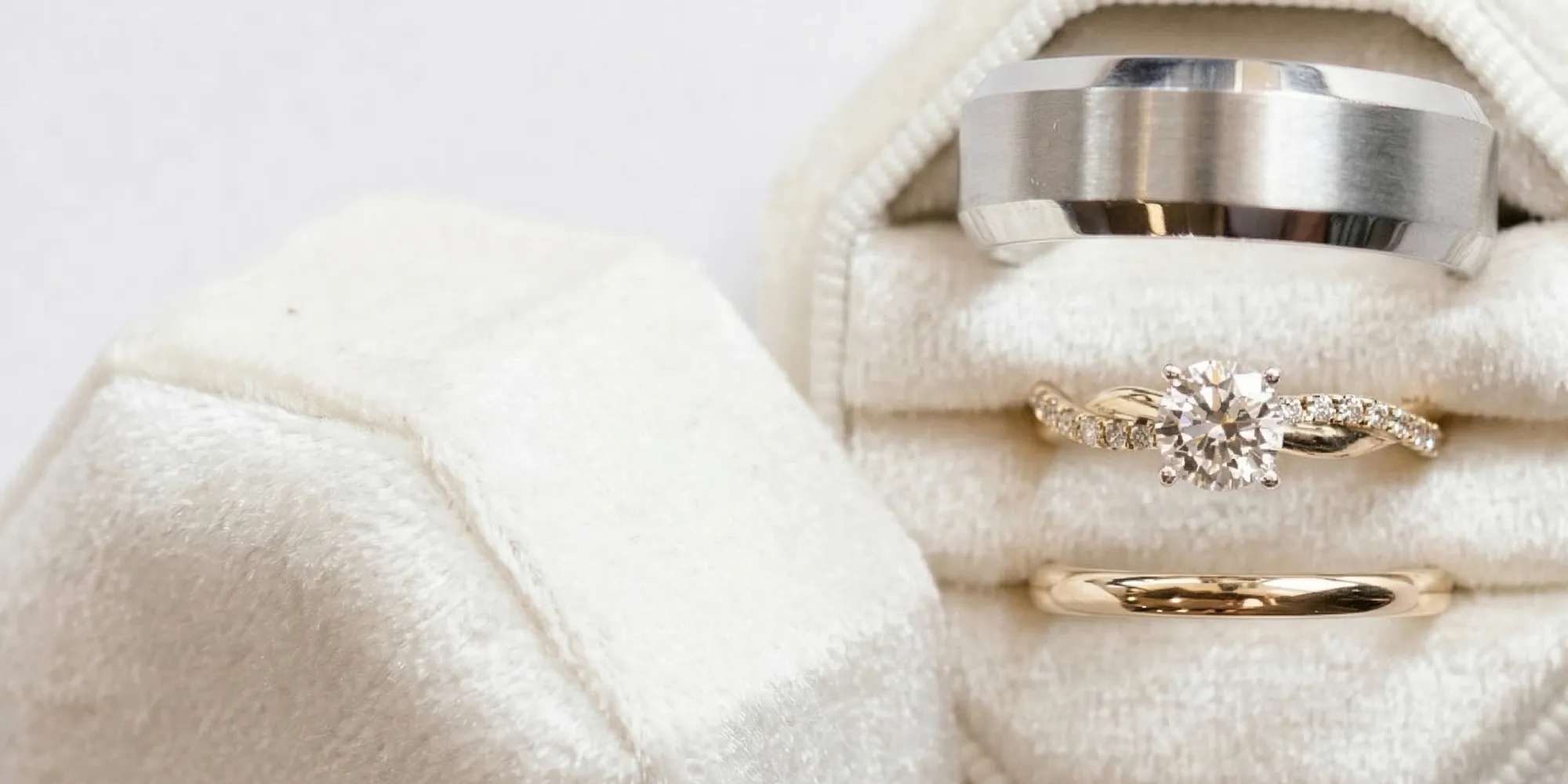 Three rings in a white velvet box: silver band, diamond engagement ring, and gold band.