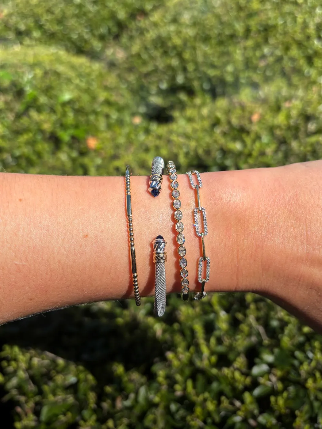 Four elegant diamond bracelets on a wrist, set against a green leafy background.