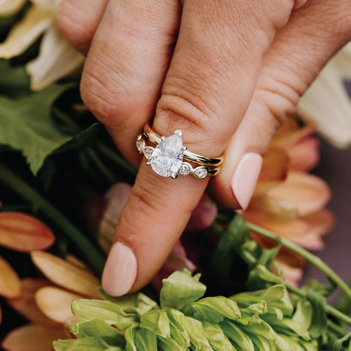 A hand wearing a gold and diamond engagement ring with a pink background.