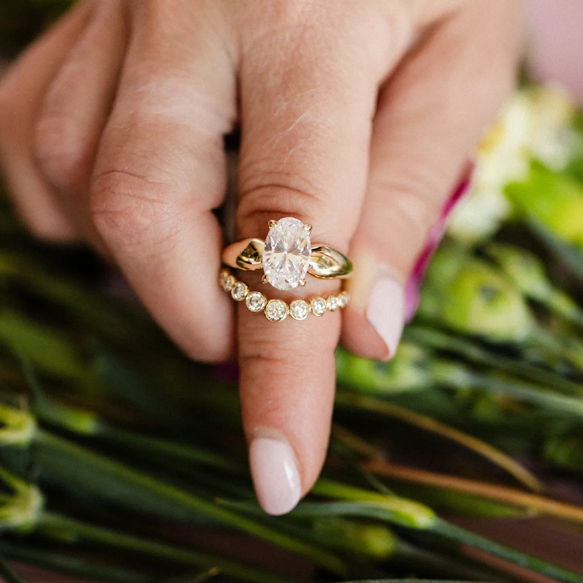 A hand with an oval diamond ring and curved diamond band over a floral background. Click for more info on the trunk show.