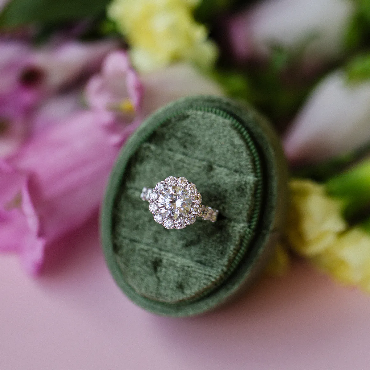 Diamond ring in a green velvet box with blurred floral background.