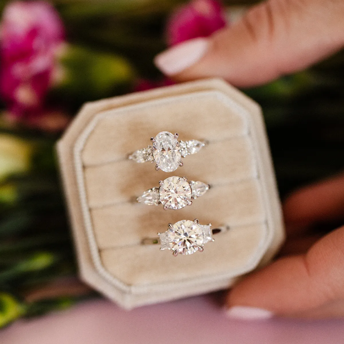 Three diamond rings displayed in a beige jewelry box with a floral background.