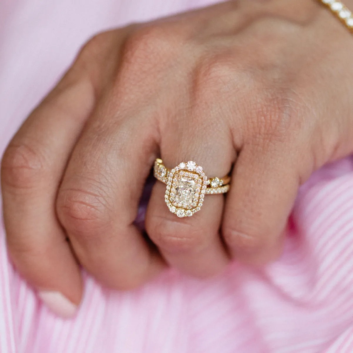A hand wearing a gold and diamond engagement ring with a pink background.