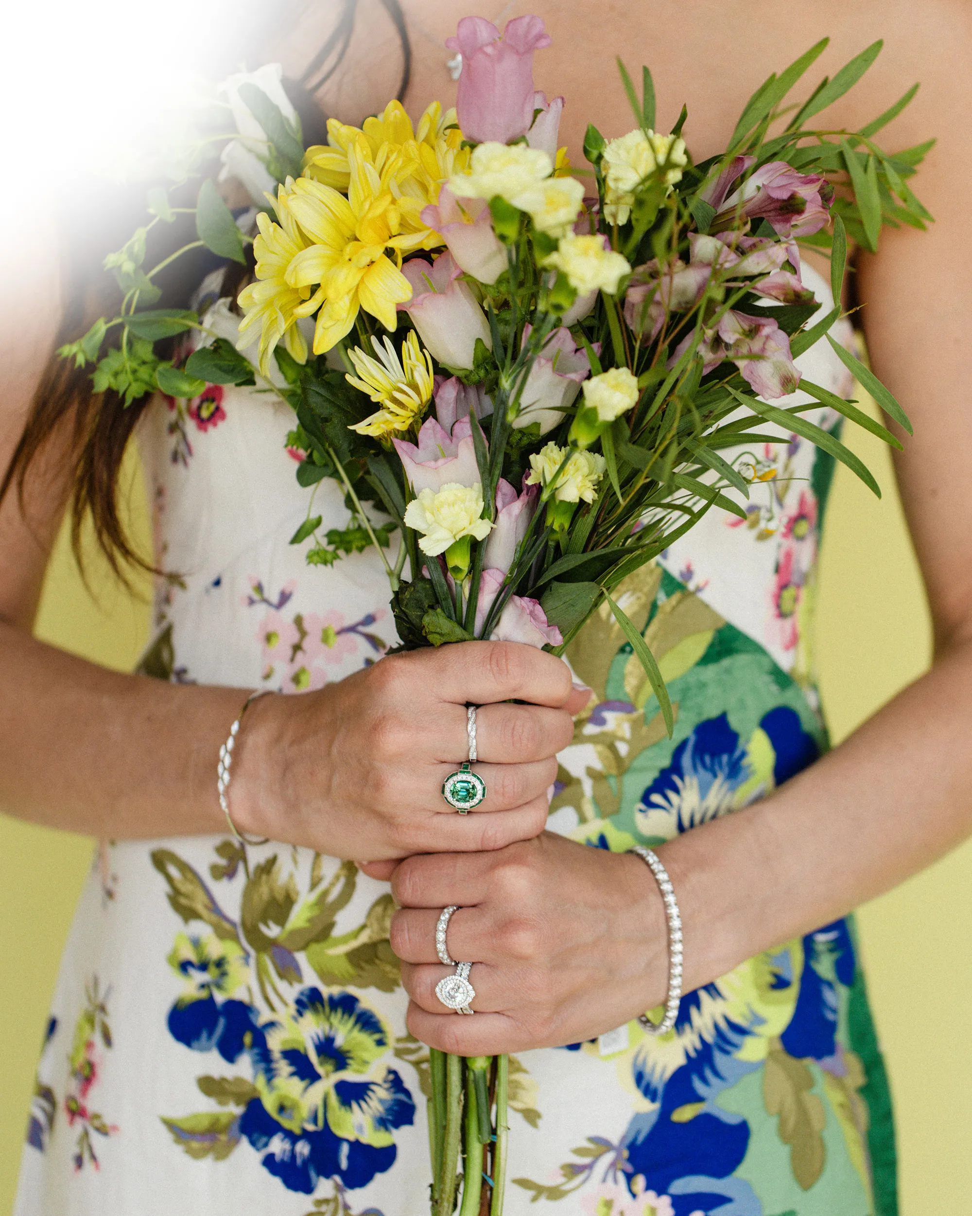 A woman in a floral dress holds a bouquet, showcasing diamond rings and a bracelet.