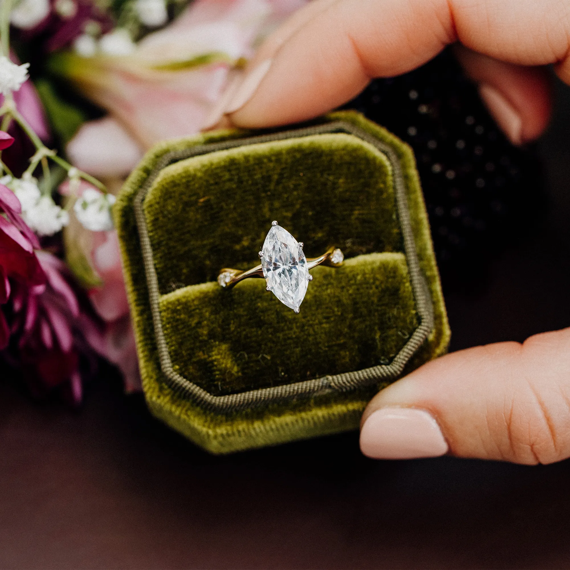 A hand with an oval diamond ring and curved diamond band over a floral background. Click for more info on the trunk show.