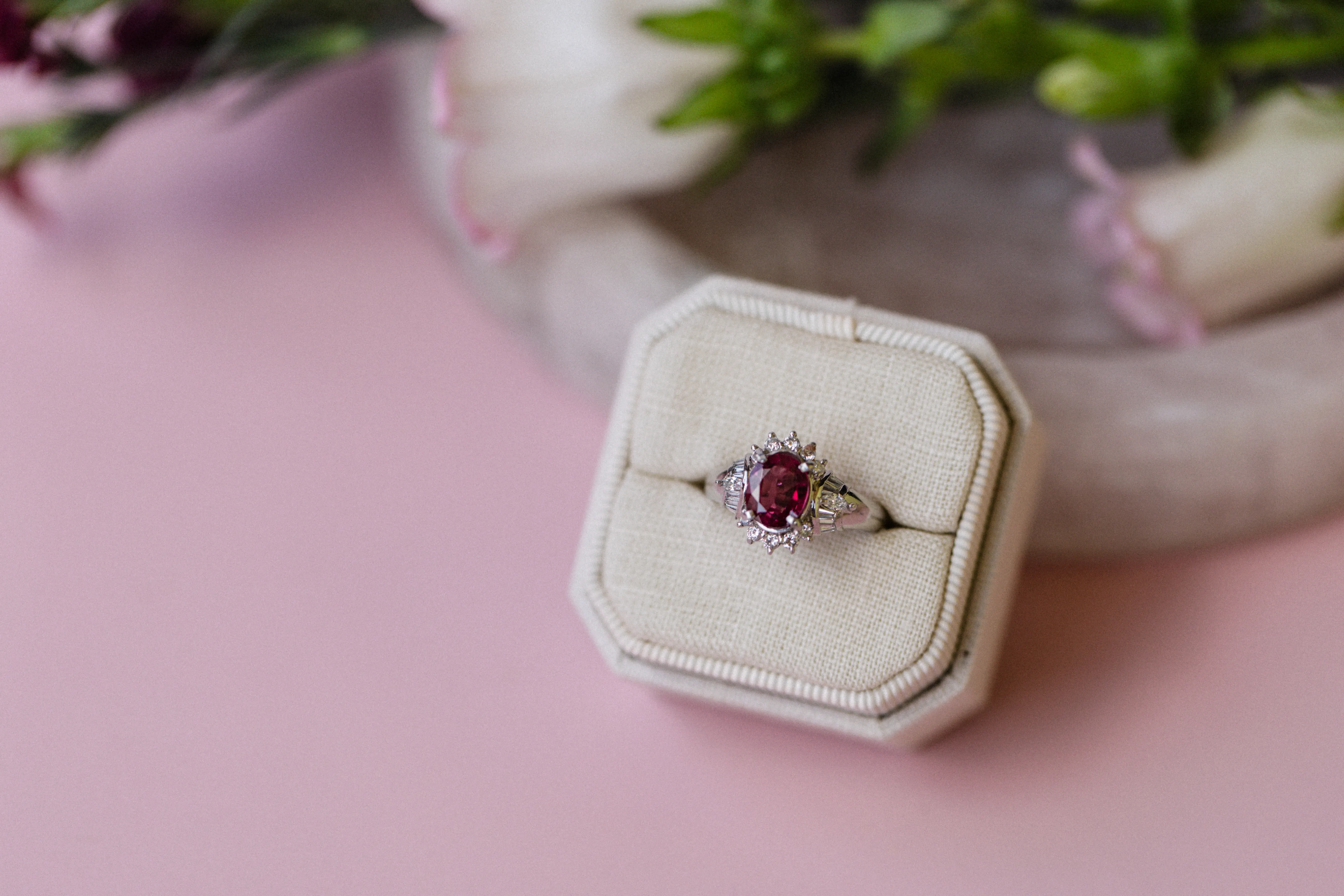 A ruby and diamond ring displayed in a beige ring box on a pink surface.