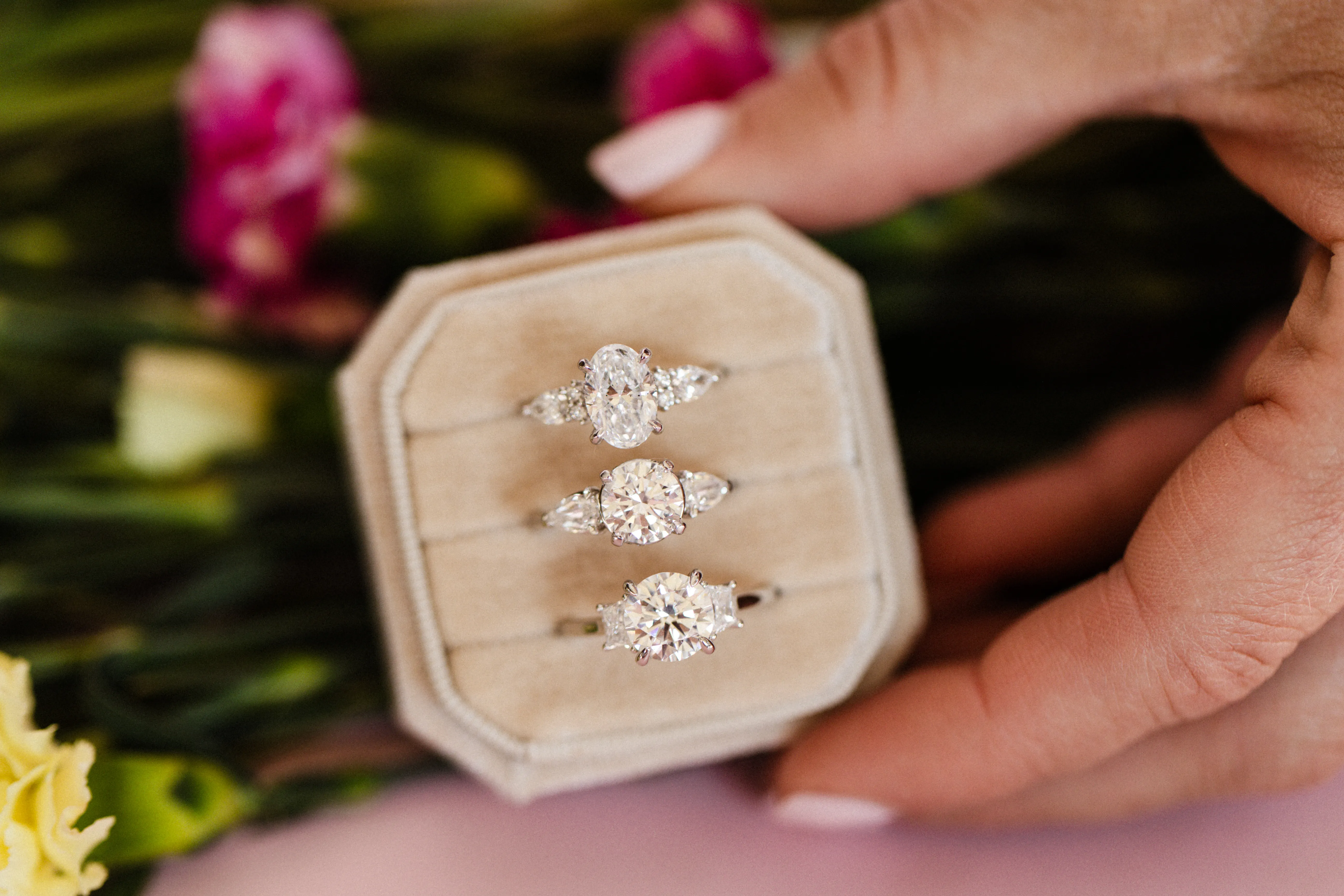 Elegant diamond engagement rings held in a hand, surrounded by flowers. Explore engagement rings.