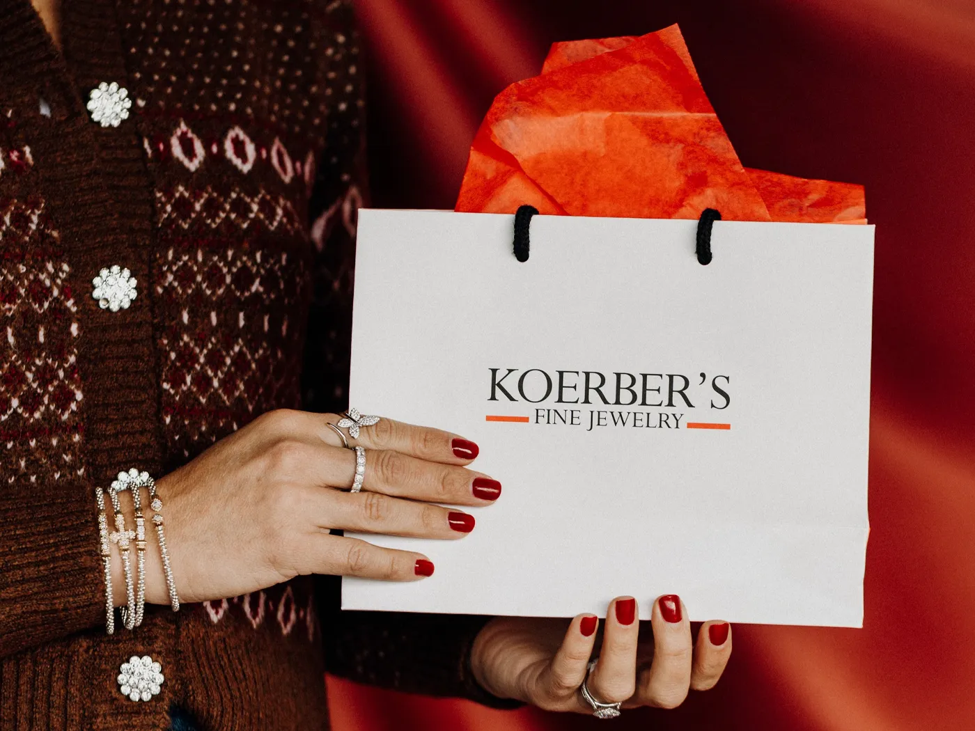 A hand holding a Koerber's Fine Jewelry bag with red tissue paper, adorned with elegant rings and bracelets.