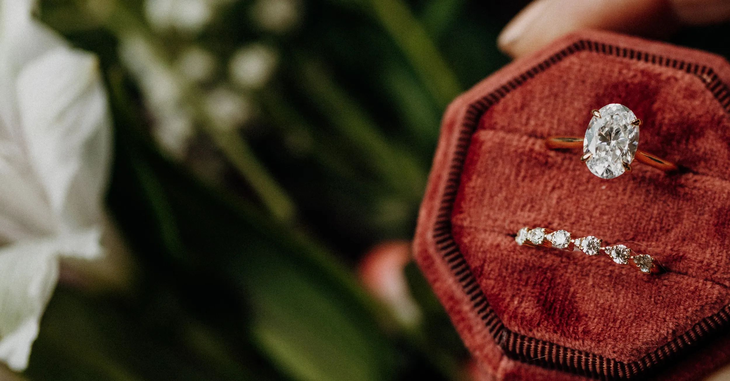 Engagement ring and wedding band displayed on a red velvet box, surrounded by greenery.