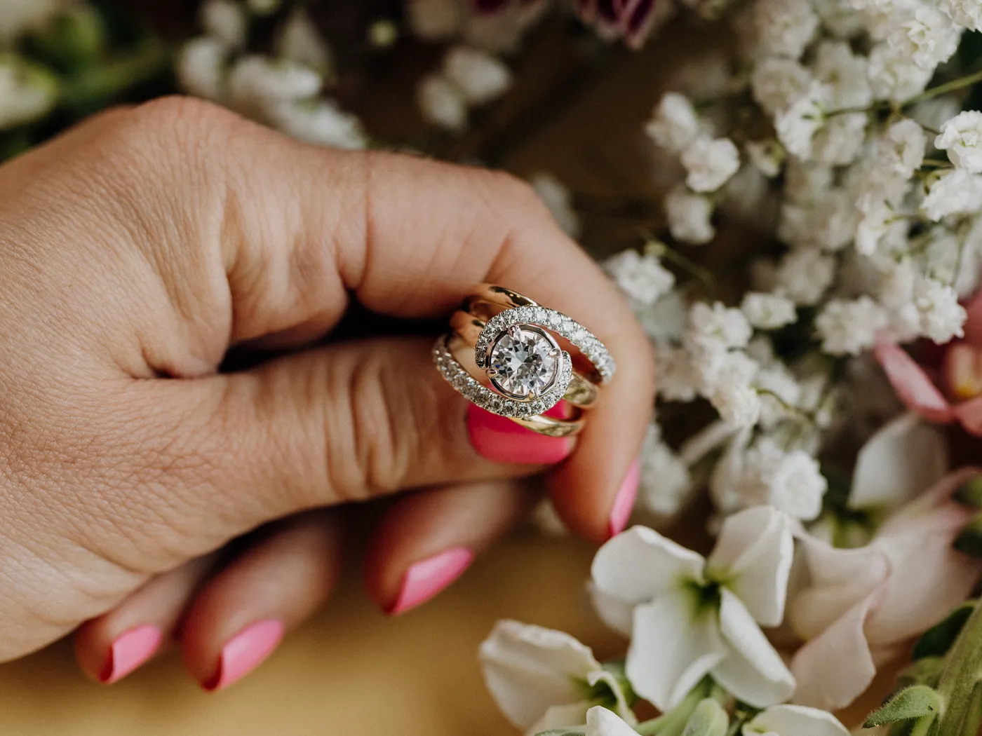 A hand holding a diamond engagement ring surrounded by flowers.