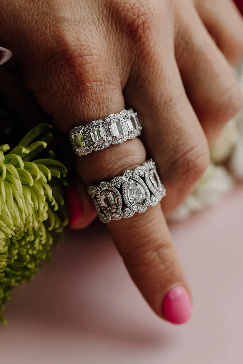 Two elegant diamond rings on a hand with pink nails, surrounded by green flowers.
