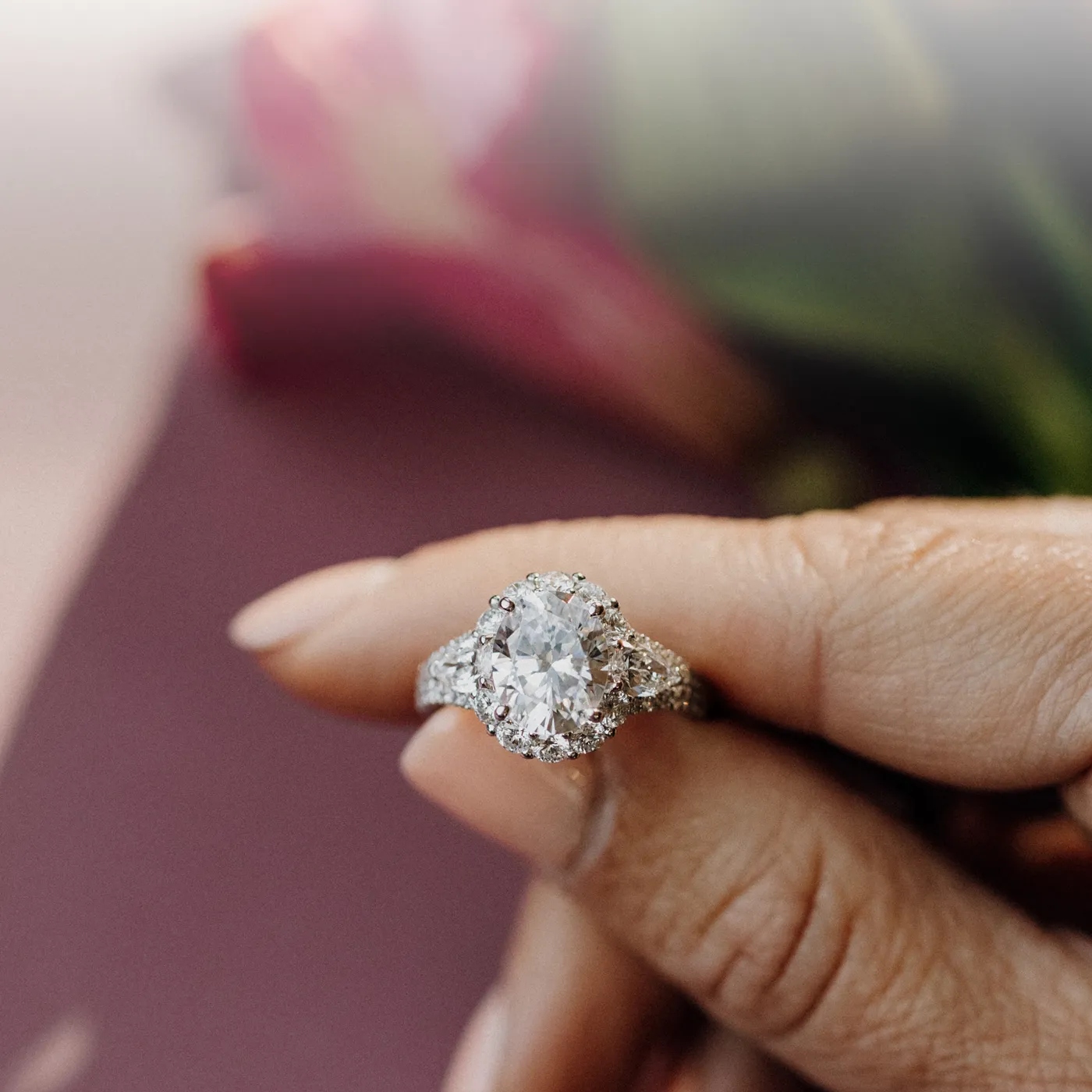 Elegant oval engagement ring with a sparkling diamond surrounded by smaller stones, set against soft floral background.