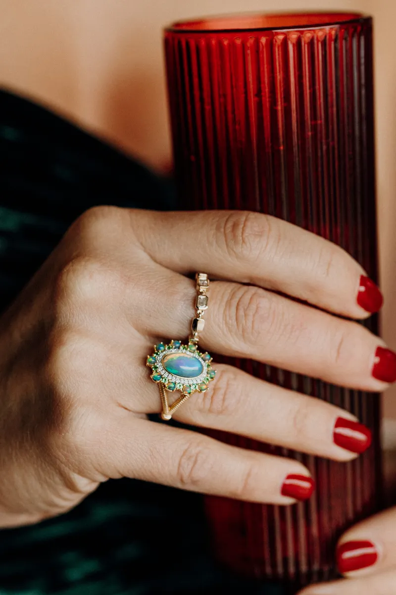 A hand with red nails holding a red glass, showcasing a gold ring with a blue stone and green accents.