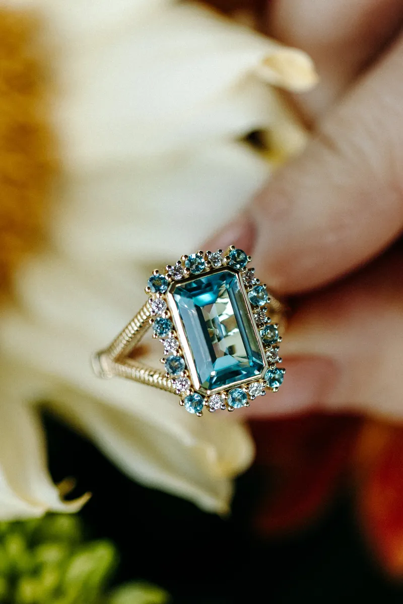 A hand holding a stunning blue gemstone ring surrounded by flowers, showcasing its intricate design and sparkle.
