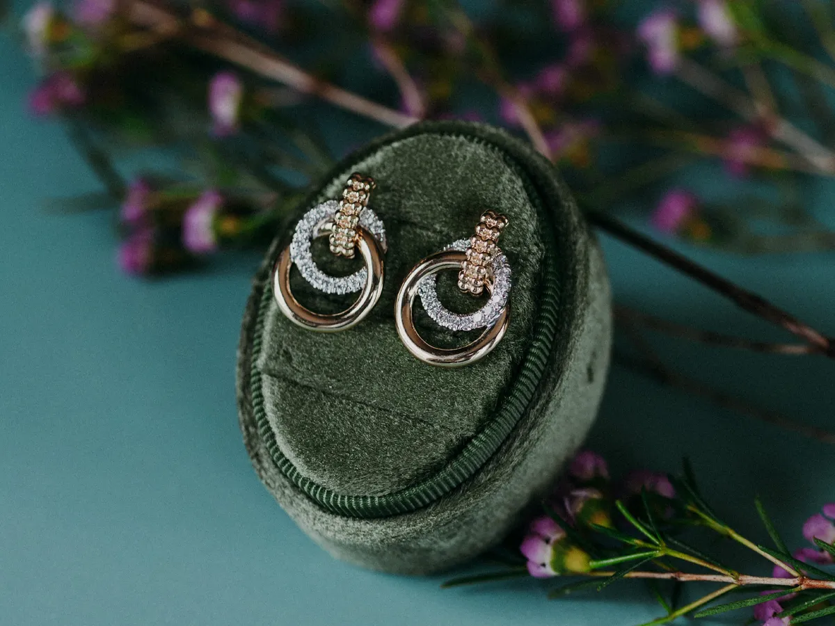 Elegant gold and silver earrings displayed in a green velvet box with flowers in the background.