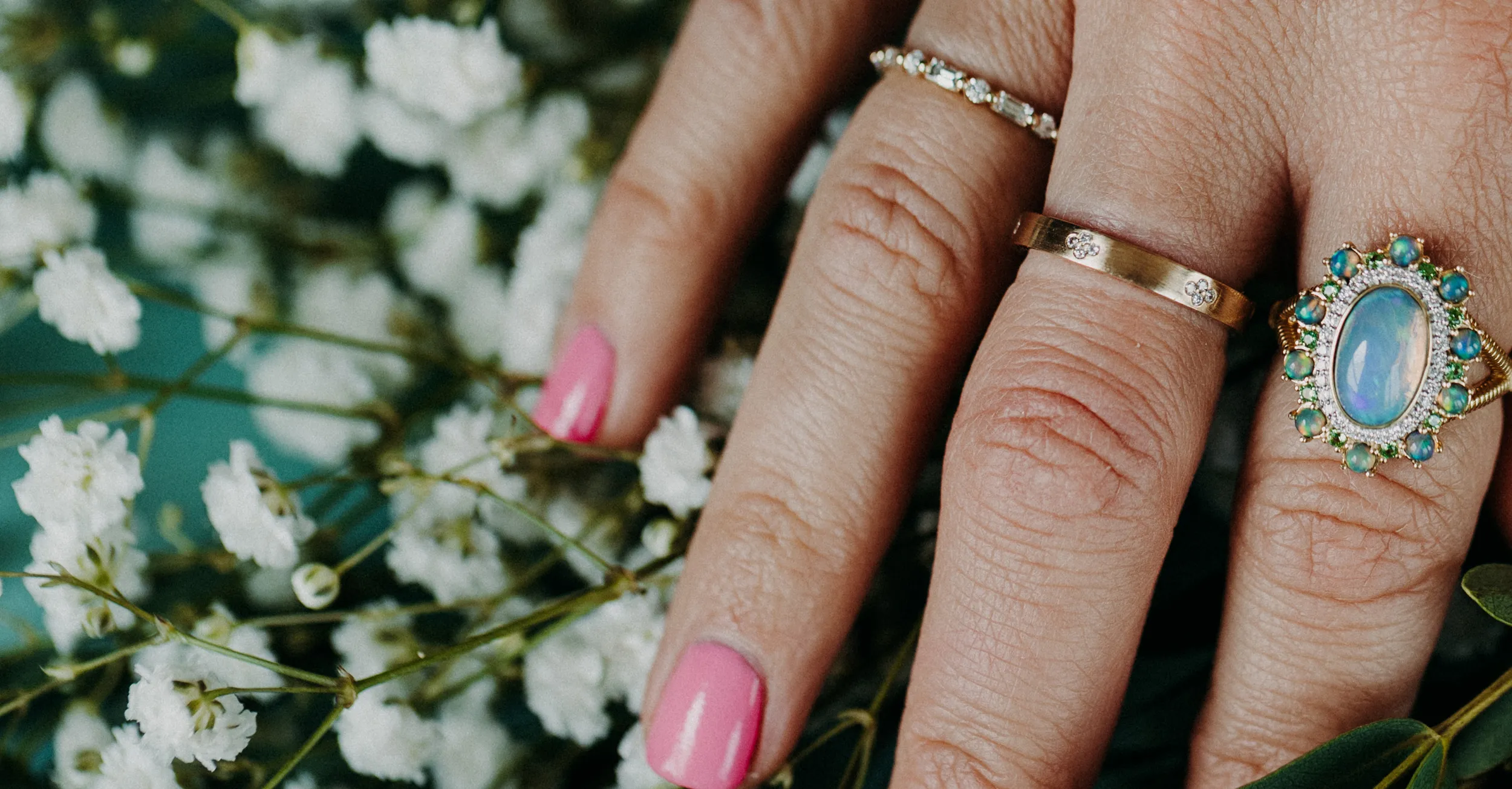 Close-up of a hand with pink nails wearing three rings, surrounded by delicate white flowers. Click to make an appointment.