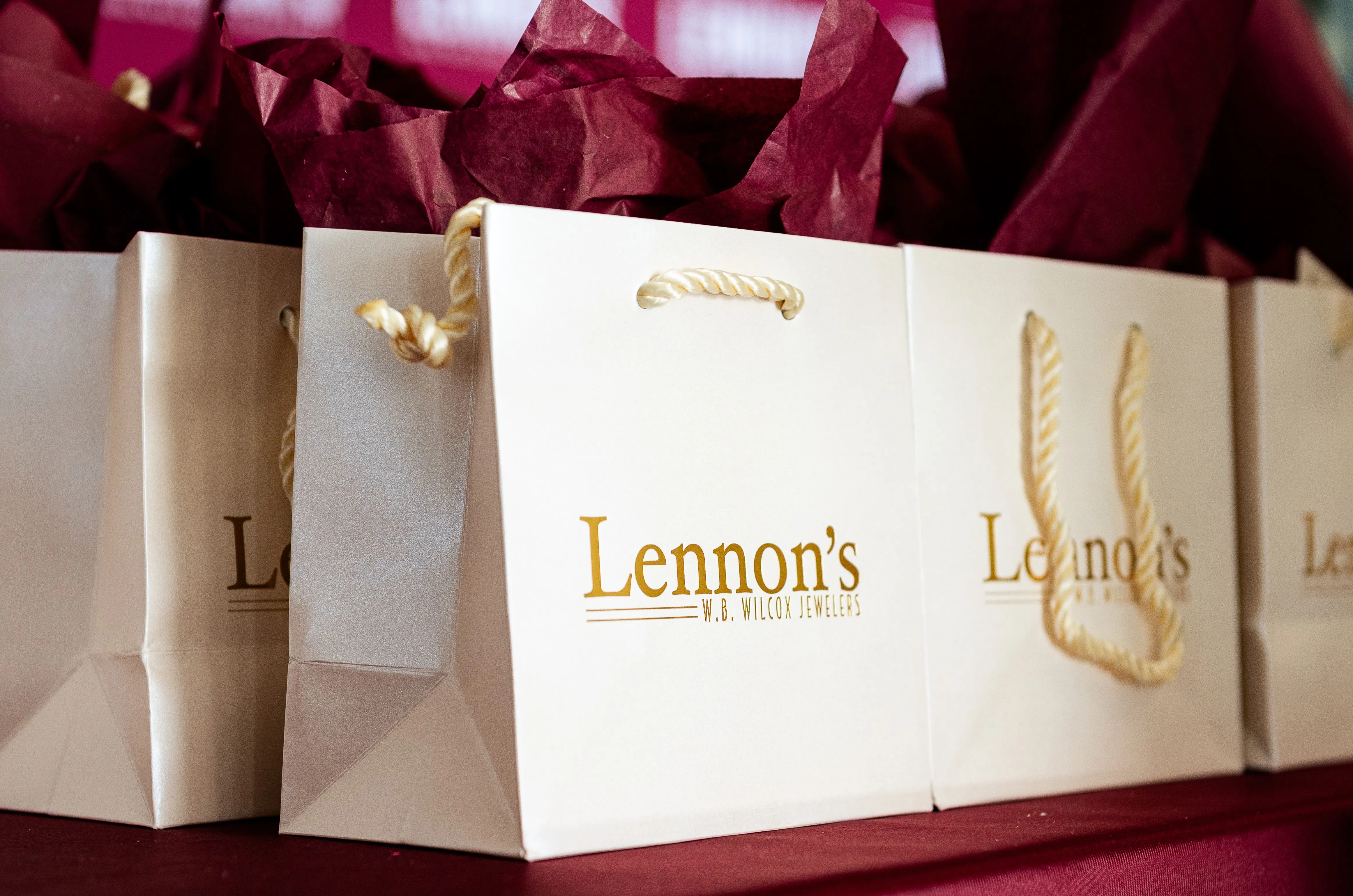 Hand with red nails wearing diamond rings, surrounded by pink and white flowers. Learn More.