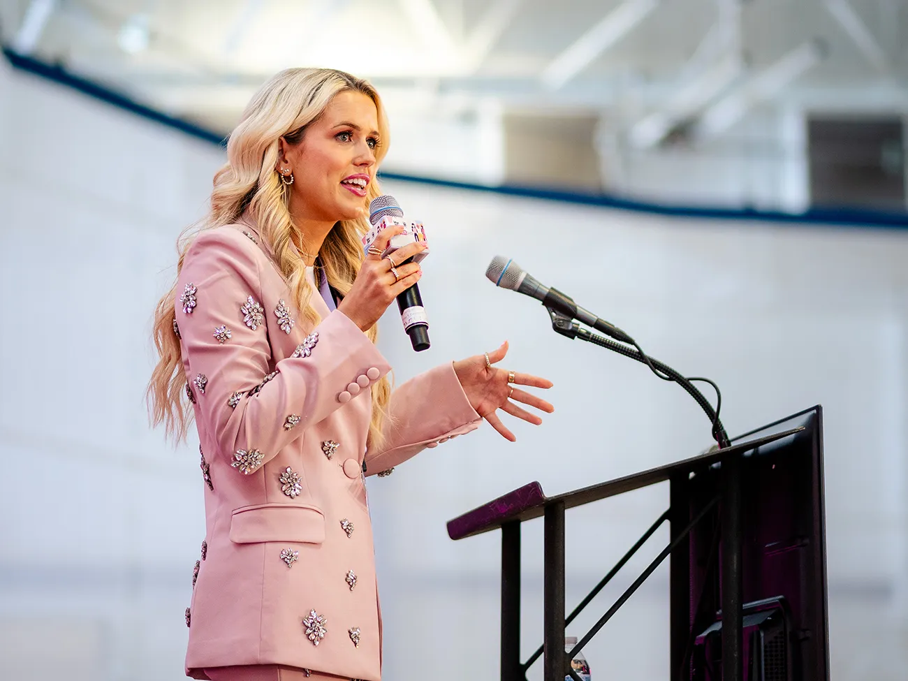 A woman in a pink embellished suit speaks at a podium with a microphone in hand.