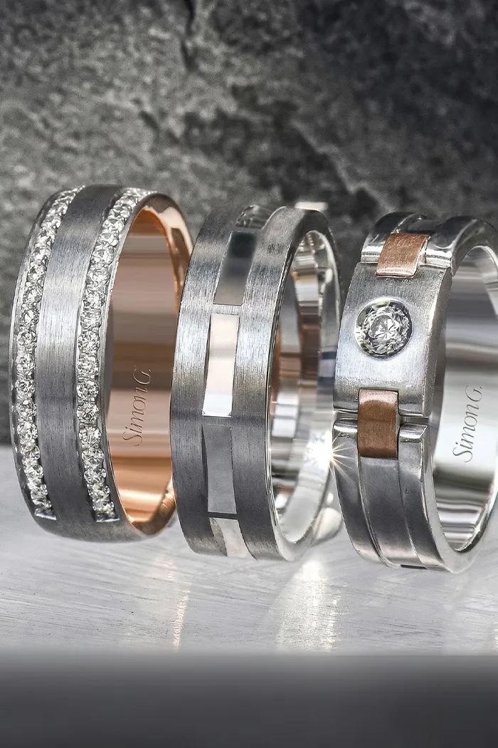 Three silver and rose gold men's wedding bands with diamond accents on a textured surface.