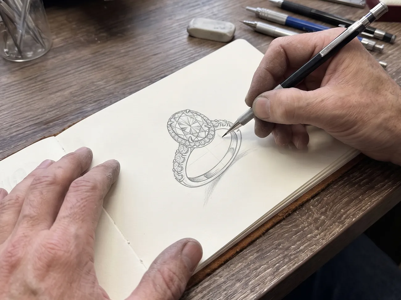 A hand sketches a detailed diamond ring design in a notebook on a wooden table.