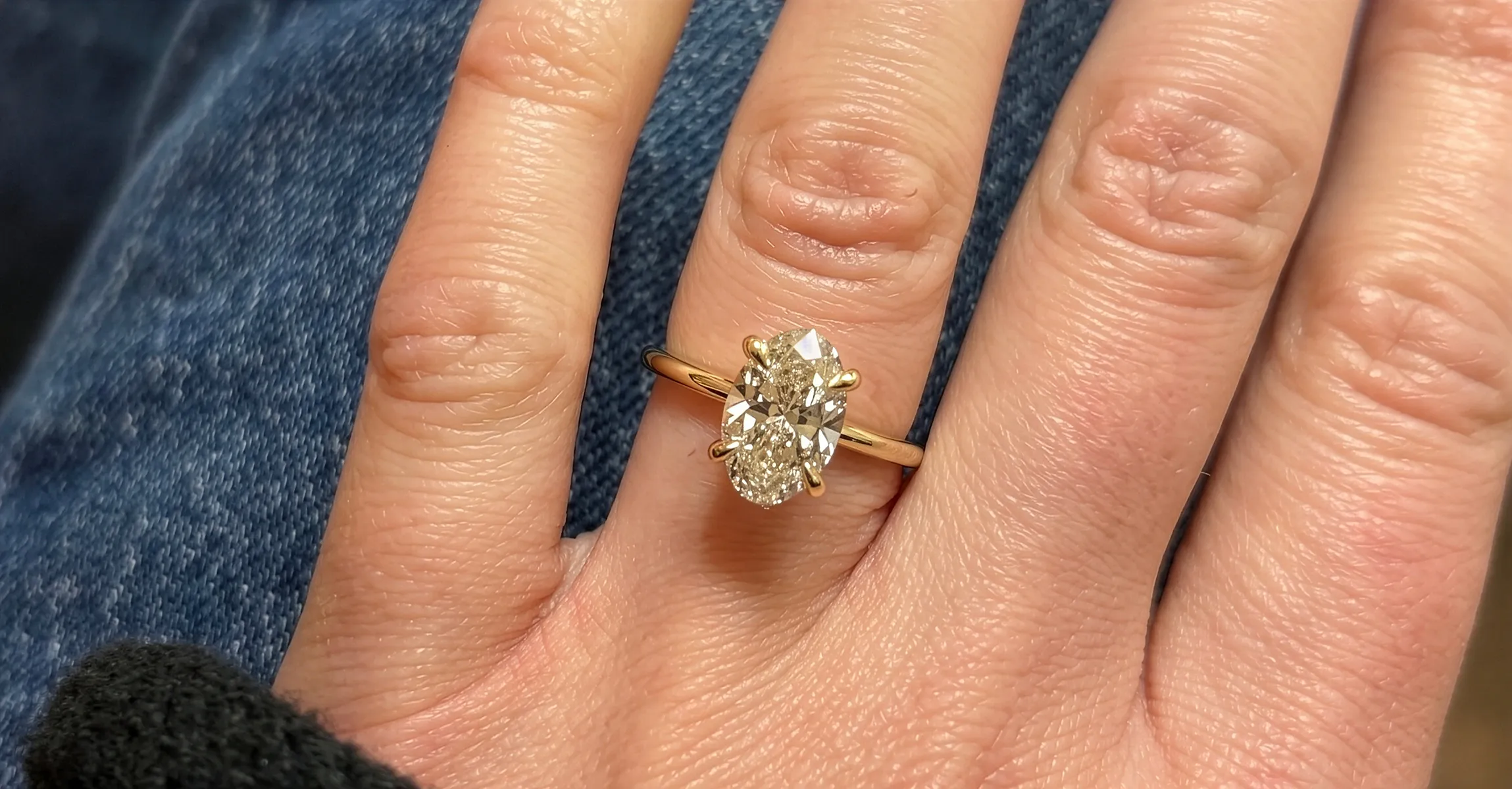 A hand wearing a gold ring with an oval diamond on a denim background. Click to view the gallery.