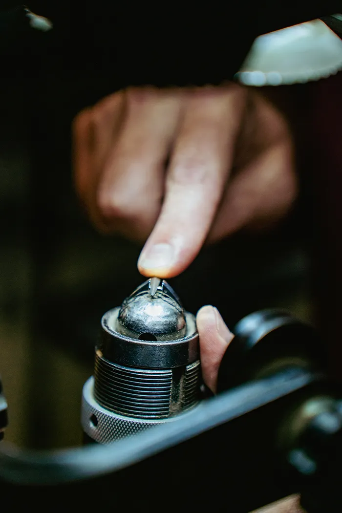 A person carefully inspects a metal piece on a jeweler's tool.