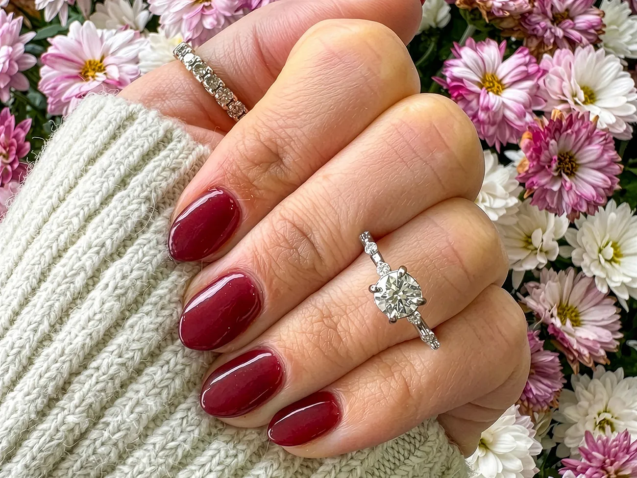 Hand with red nails wearing diamond rings, surrounded by pink and white flowers. Learn More.