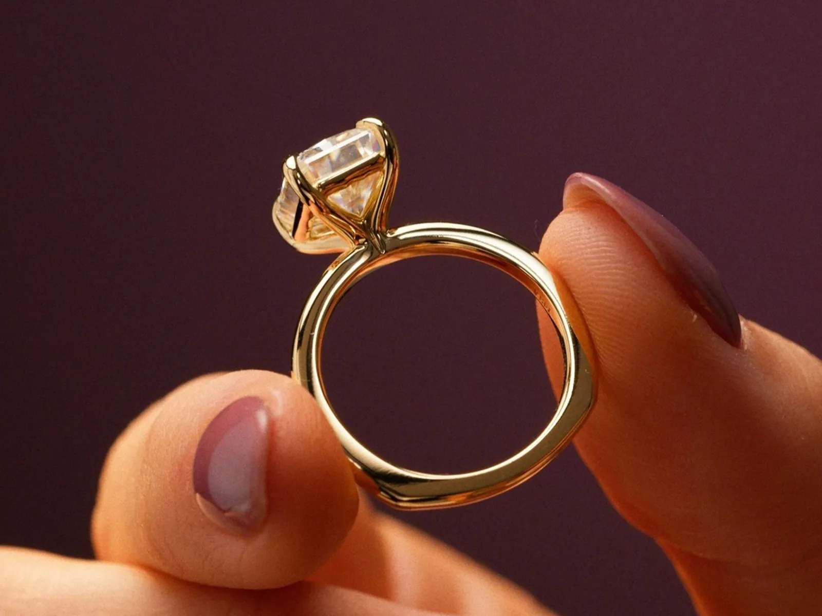 A hand holding a gold engagement ring with a large diamond against a purple background.