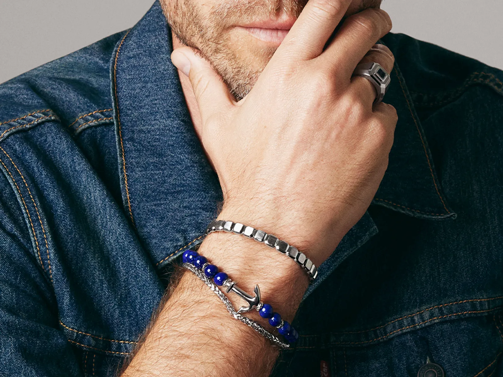 Man in denim jacket wearing silver and blue beaded bracelets, touching face.