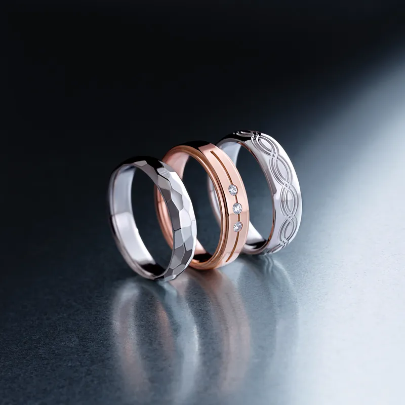 Three elegant men's wedding bands in silver, rose gold, and patterned white gold displayed on a reflective surface.