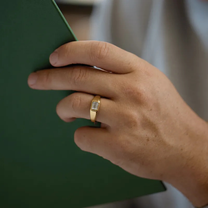 A hand holding a green folder, wearing a gold men's engagement ring with a rectangular stone.