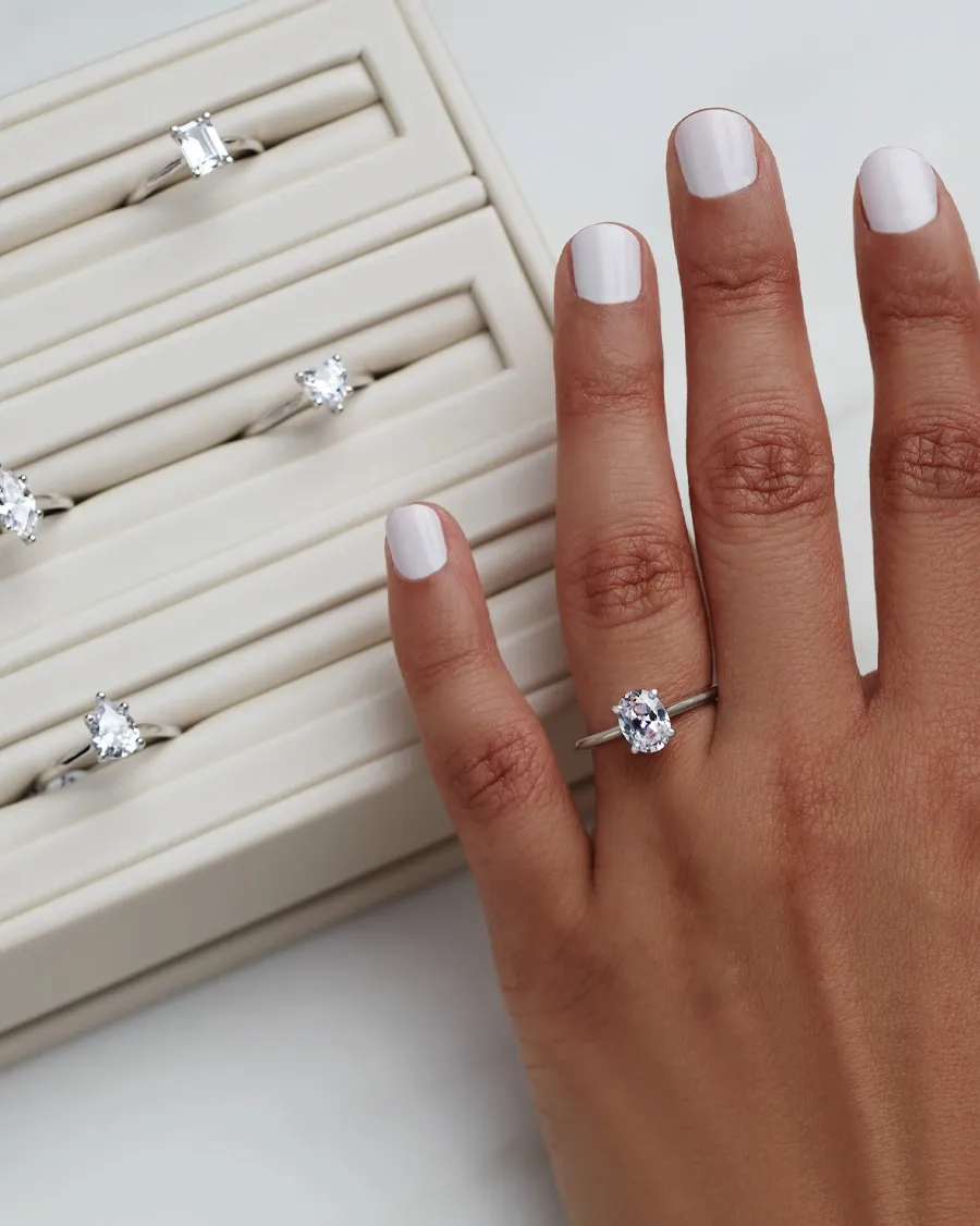 A hand with a diamond ring showcases various engagement rings in a display case.