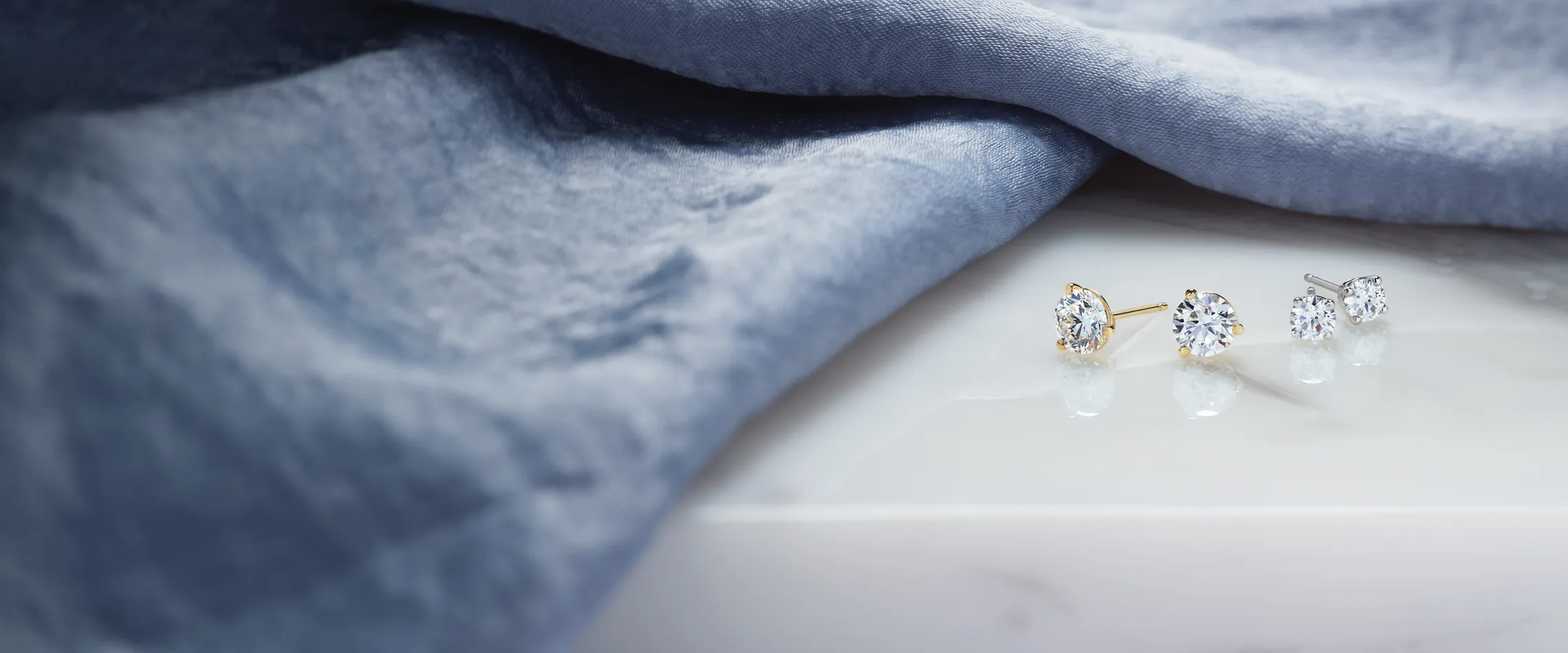 Elegant diamond stud earrings on a marble surface, draped with soft blue fabric.