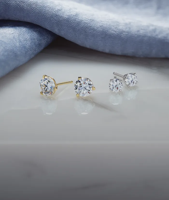 Elegant diamond stud earrings on a marble surface, draped with soft blue fabric.