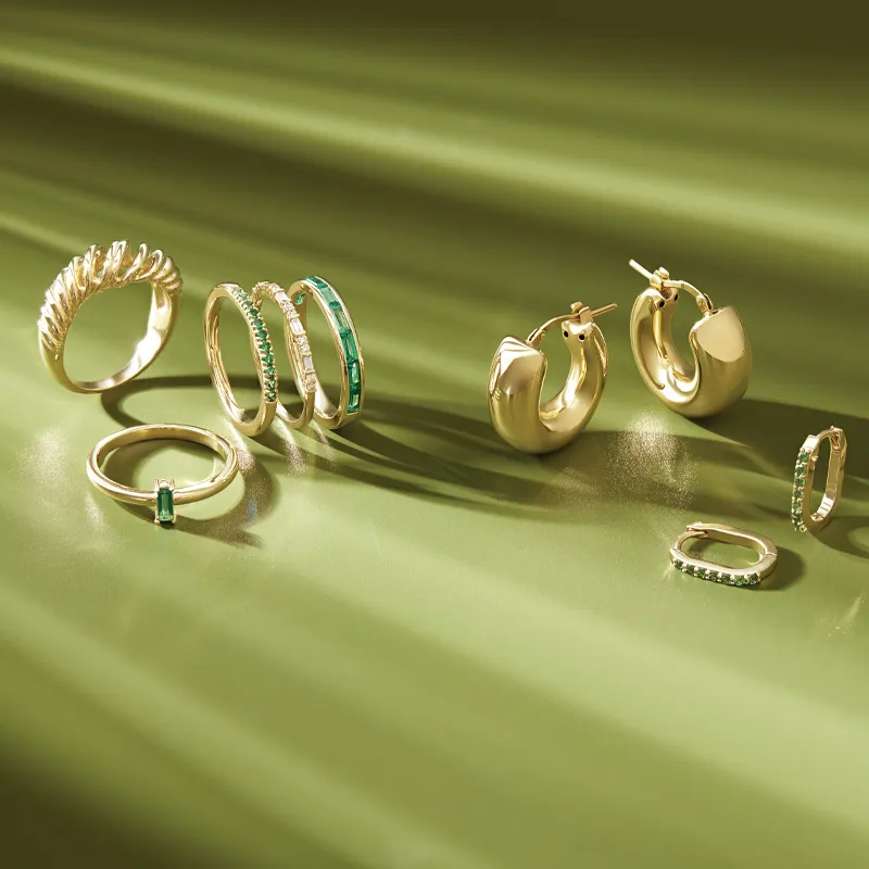 Gold jewelry collection featuring rings and earrings with emerald accents, displayed on a green background.
